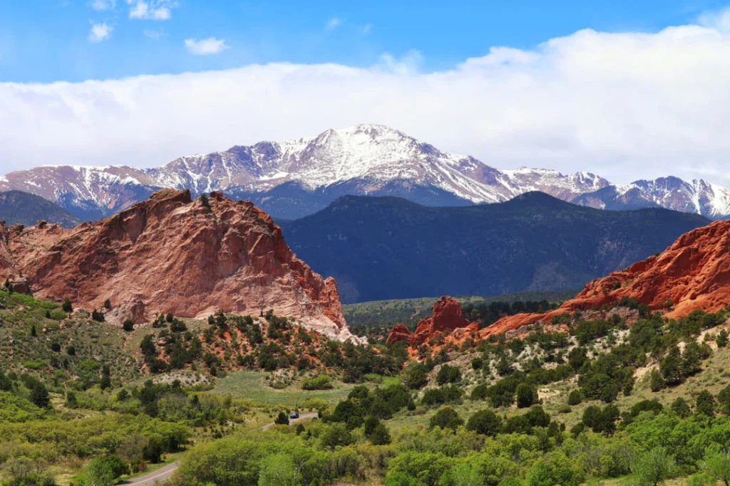 Rode rotsformaties van Garden of the Gods omringd door groene begroeiing met de besneeuwde top van Pikes Peak in de verte onder een blauwe lucht