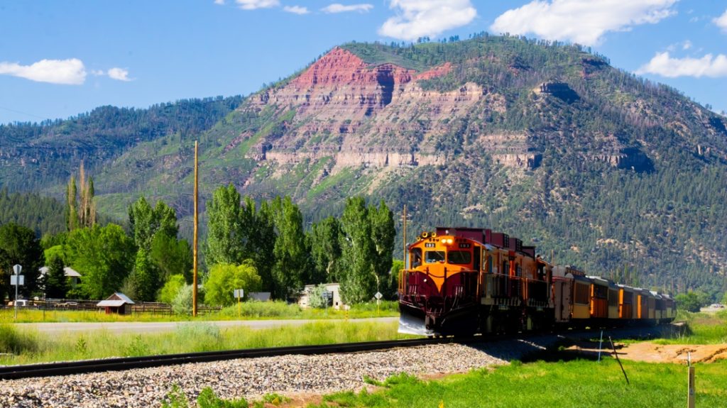 Oranje locomotief met wagons rijdt over een spoor voor een gestreepte rode en groene berg in Colorado