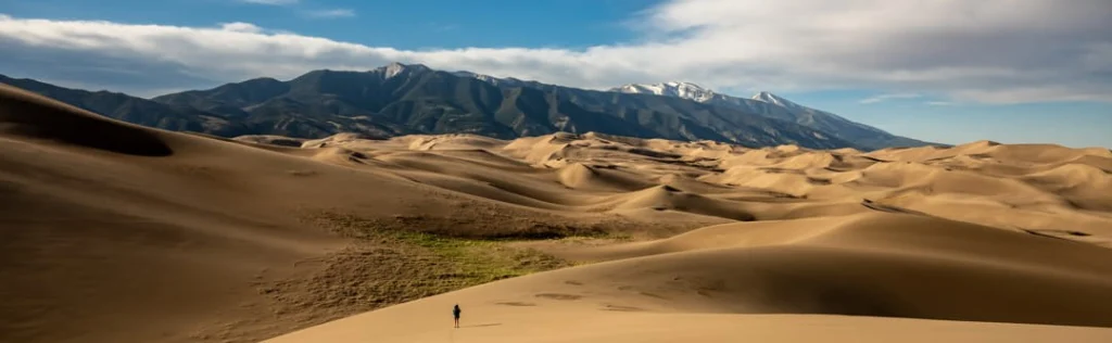 Een kleine wandelaar loopt tussen de uitgestrekte zandduinen van Great Sand Dunes National Park in Colorado, met besneeuwde bergen op de achtergrond