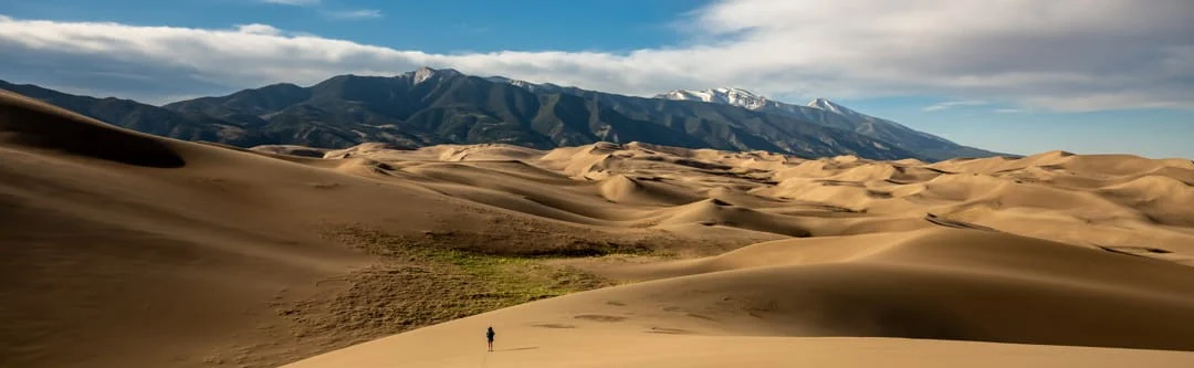 Een kleine wandelaar loopt tussen de uitgestrekte zandduinen van Great Sand Dunes National Park in Colorado, met besneeuwde bergen op de achtergrond
