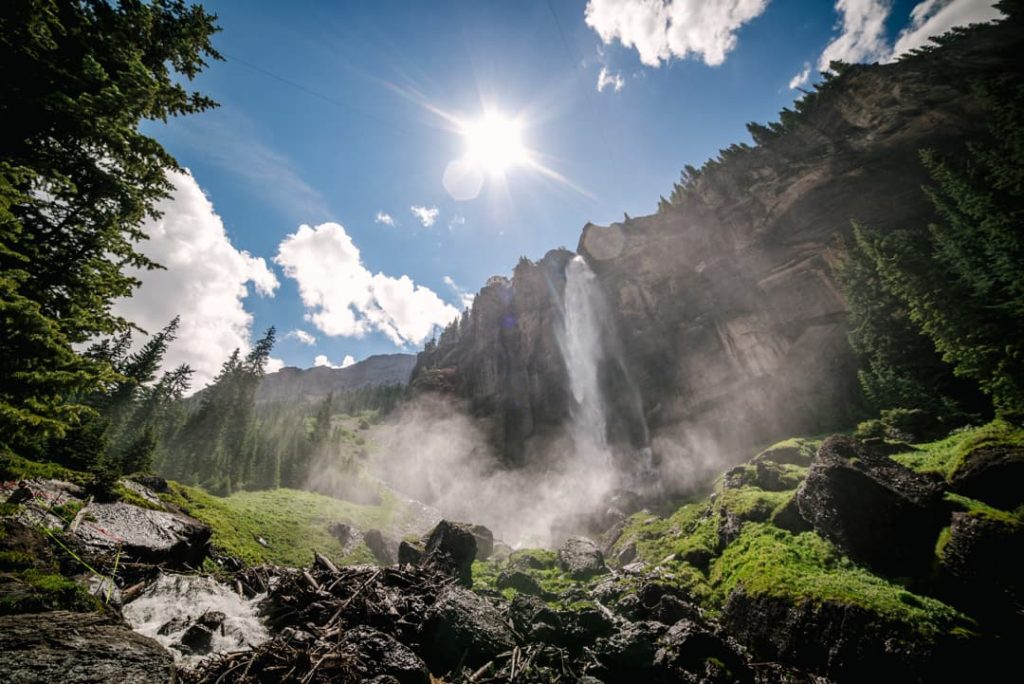 Waterval stroomt langs een rotswand omringd door dennenbomen en mos met de zon fel schijnend boven de bergen in Colorado