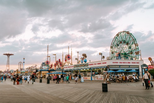 Drukke houten boardwalk van Coney Island met kraampjes, een reuzenrad en wandelaars onder een bewolkte lucht bij zonsondergang