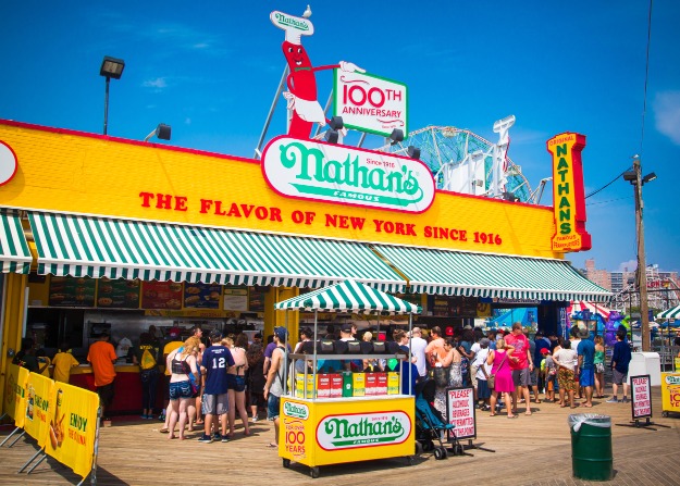 Drukbezochte hotdogkraam Nathan's Famous met geel groene luifel en wachtende mensen op de boardwalk van Coney Island