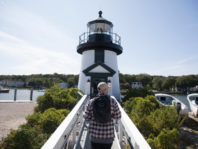 Bezoeker loopt over een witte loopbrug naar een zwart witte vuurtoren met boten en groen op de achtergrond in Connecticut
