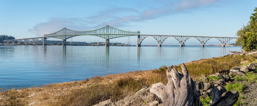Lange groene stalen boogbrug die over kalm water loopt met op de voorgrond gras en verweerd drijfhout