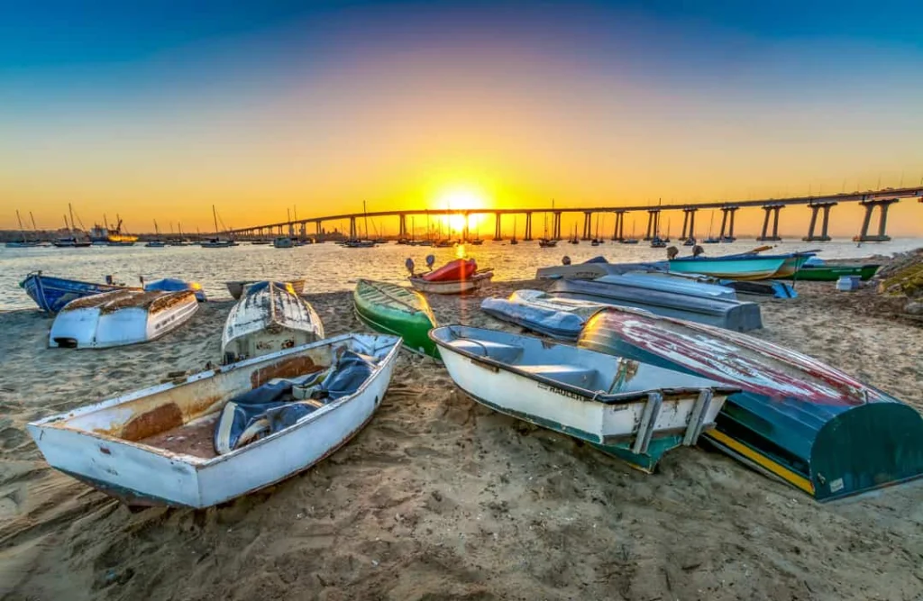 Verweerde roeibootjes liggen op het zand bij zonsopgang met de lange Coronado Bridge en zeilboten op de achtergrond