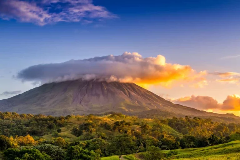 De conische Arenal-vulkaan in Costa Rica steekt boven groen regenwoud uit terwijl de avondzon de wolken oranje kleurt