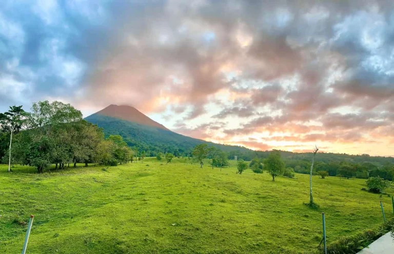 Groene vlakte met bomen en op de achtergrond de Arenal vulkaan onder een kleurrijke bewolkte lucht bij zonsondergang