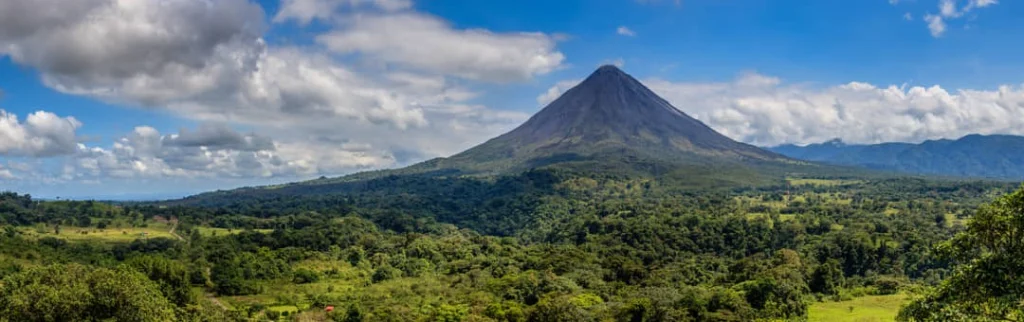 Symmetrische kegelvormige vulkaan met een lichte rooksluier boven groen regenwoud en landbouwgrond onder een blauwe wolkenlucht