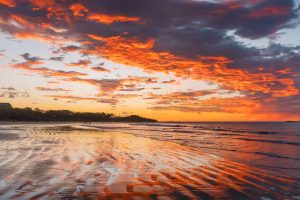 Fel oranje en roze wolkenlucht weerspiegelt in het natte zand van een leeg strand bij zonsondergang