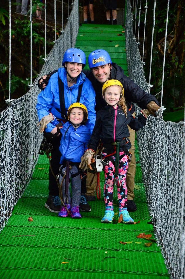 Een gezin met blauwe en gele helmen en klimtuig staat glimlachend op een groene hangbrug in het regenwoud tijdens een ziplineavontuur