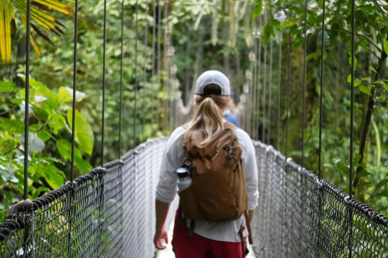 Persoon met rugzak en pet loopt over een hangende voetbrug omgeven door dicht groen tropisch regenwoud in Costa Rica