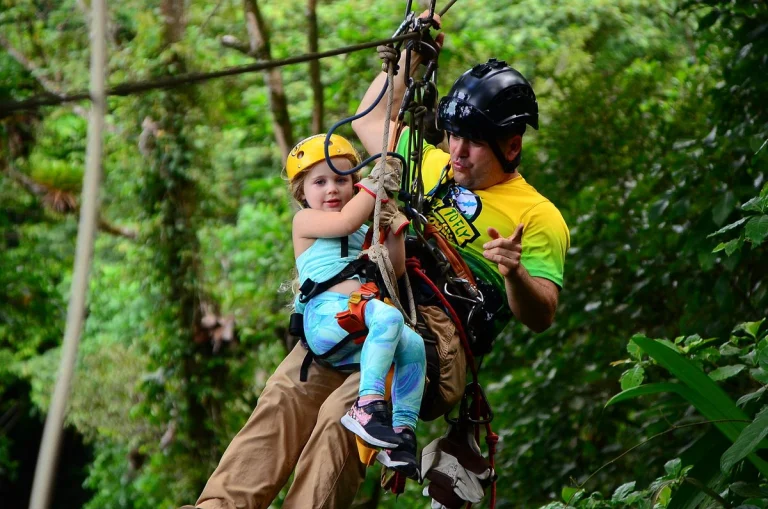 Een jonge kind met gele helm hangt aan een zipline in het regenwoud terwijl een gids haar vasthoudt en begeleidt
