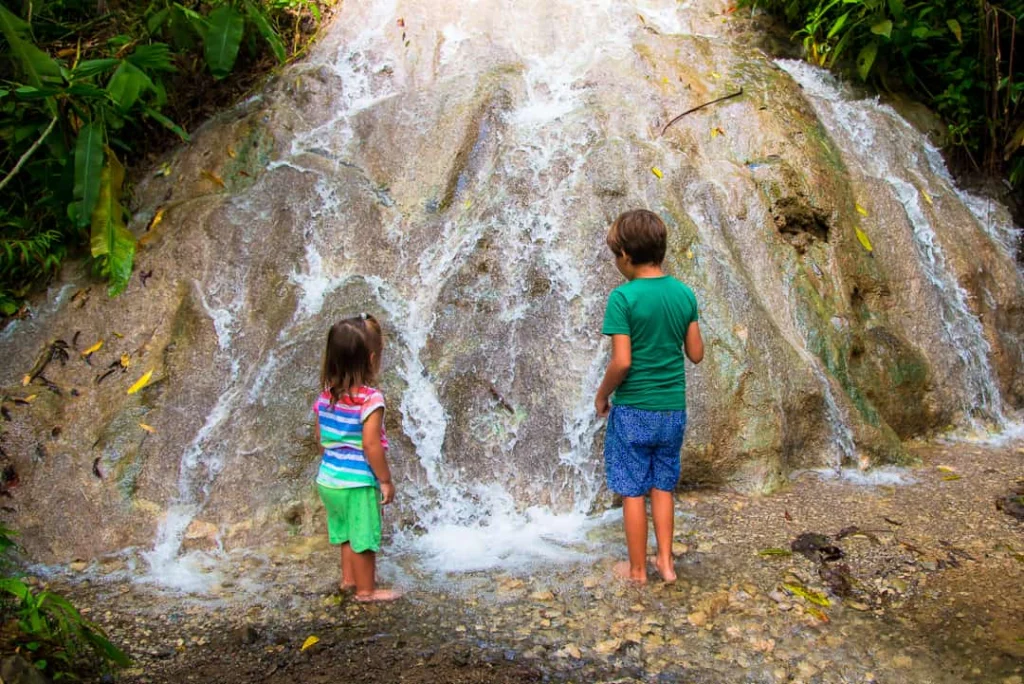 Twee kinderen staan met hun voeten in het water aan de voet van een brede waterval over rotsen in het regenwoud