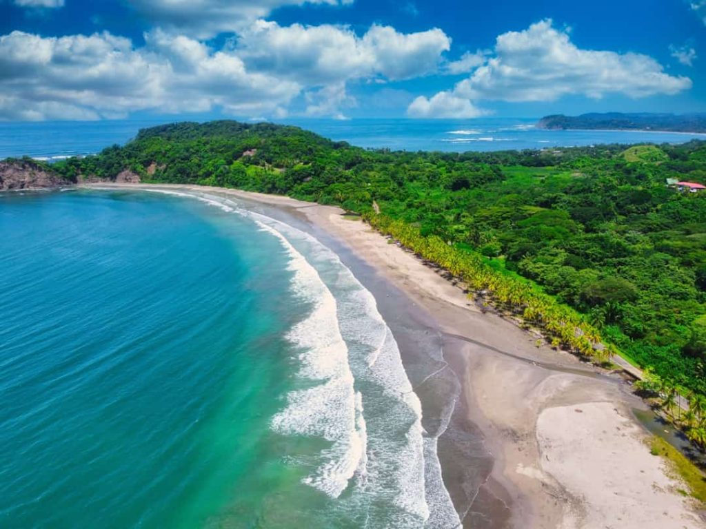 Luchtfoto van een halvemaanvormige zandbaai met turkooize golven en een groen beboste landtong aan de Pacifische kust van Costa Rica