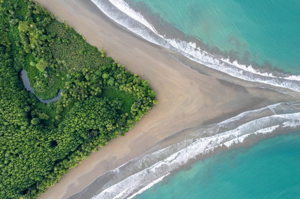 Luchtfoto van een driehoekige groene mangrovepunt tussen twee zandstranden met turkooize golven aan weerszijden in Costa Rica
