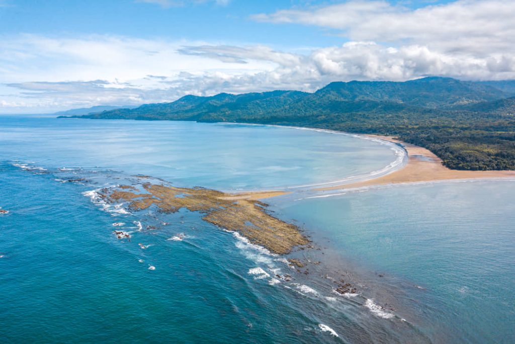 Luchtfoto van een lange baai met een rotsachtig rif in zee en een beboste bergketen aan de horizon in Costa Rica