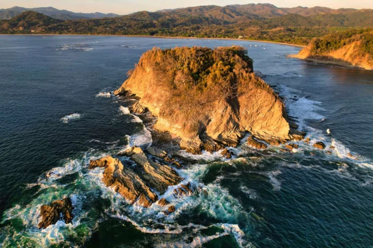Luchtfoto van een steile met bos begroeide rotspunt omringd door schuimende golven in de Stille Oceaan met een baai op de achtergrond