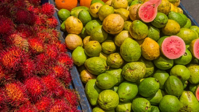 Bakken vol felrode rambutans en groengele guaves op een markt, met een enkele doorgesneden roze guave ertussen