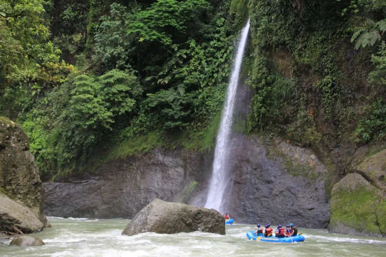 Een groep raften in een blauwe boot over de wilde Pacuare River terwijl op de achtergrond een hoge waterval tussen weelderig groen naar beneden stort