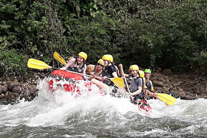 Groep raftende reizigers met helmen en zwemvesten peddelt over een minder woelig stuk rivier tussen dicht regenwoud in Costa Rica