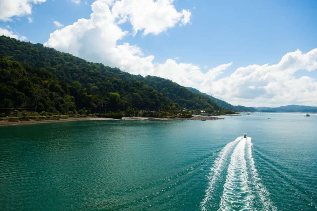 Een speedboot vaart met een schuimend kielzog over kalm turquoise water langs een kust met groene heuvels