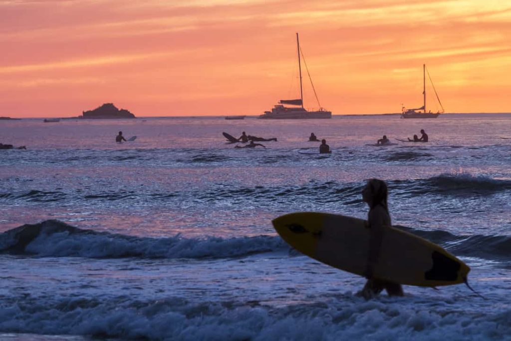 Silhouet van een surfer die met een gele surfplank uit zee loopt terwijl boten voor anker liggen bij een oranje zonsondergang