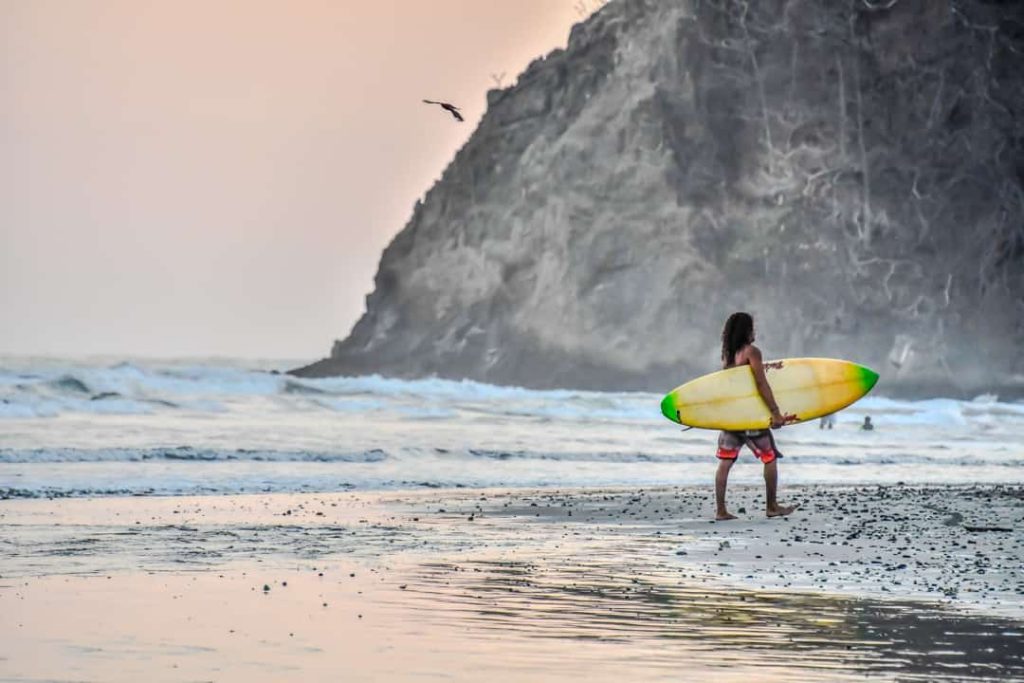 Surfer loopt met een geel surfboard onder de arm langs de branding, met een rotsachtige kaap op de achtergrond bij zonsondergang in Costa Rica