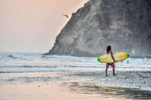 Surfer loopt met een geel surfboard onder de arm langs de branding, met een rotsachtige kaap op de achtergrond bij zonsondergang in Costa Rica