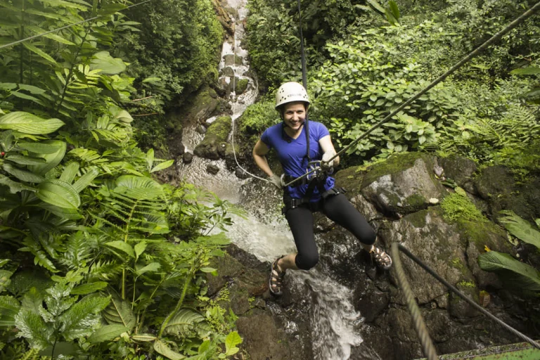 Een lachende vrouw met helm abseilt via touwen langs een waterval omgeven door weelderig groen regenwoud