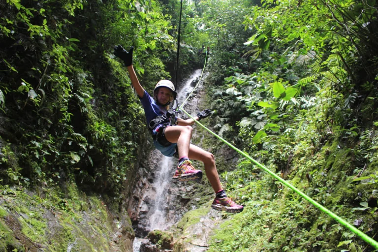 Een vrouw met helm abseilt aan een groen touw langs een waterval in een weelderige jungle omgeving