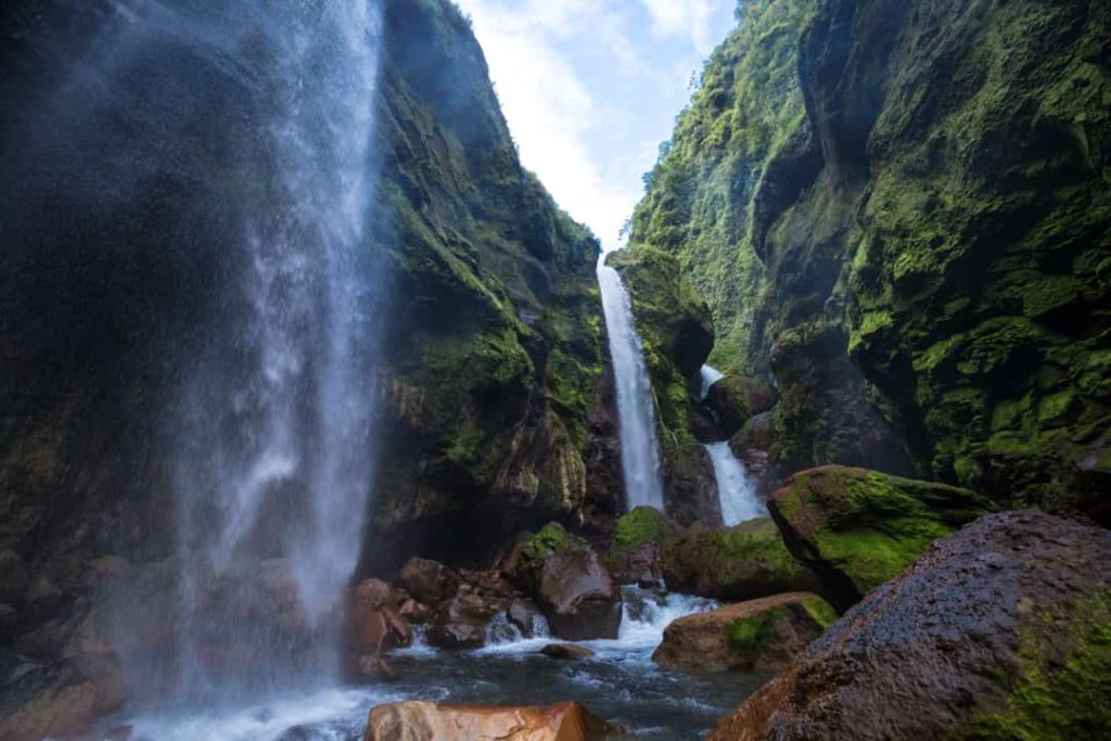 Een hoge waterval stort neer in een smalle canyon met mosbegroeide rotswanden en een tweede waterval op de achtergrond