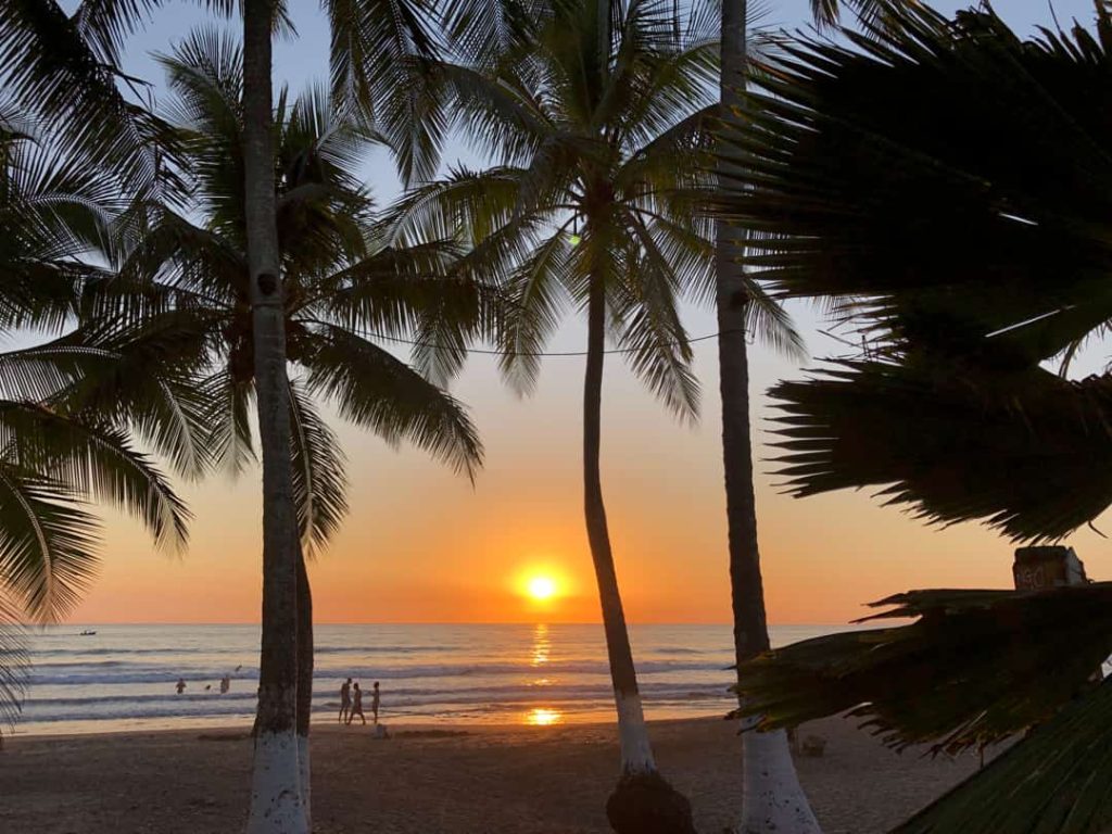 Silhouetten van palmbomen en wandelaars op het strand tegen een oranje gloeiende zonsondergang boven zee