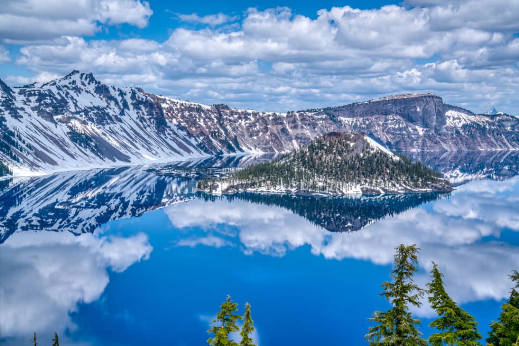 Besneeuwde bergwanden en Wizard Island weerspiegelen in het spiegelgladde blauwe water van Crater Lake onder een bewolkte lucht in Oregon