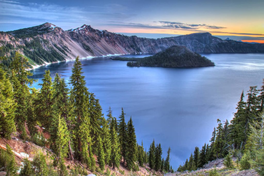 Diepblauw water van Crater Lake met Wizard Island en de steile kraterwanden onder een gekleurde avondlucht in Oregon