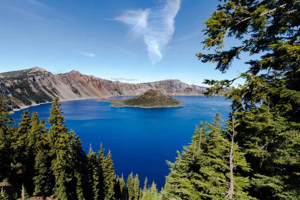 Uitzicht tussen dennentakken door op het diepblauwe water van Crater Lake met Wizard Island op een zonnige dag in Oregon