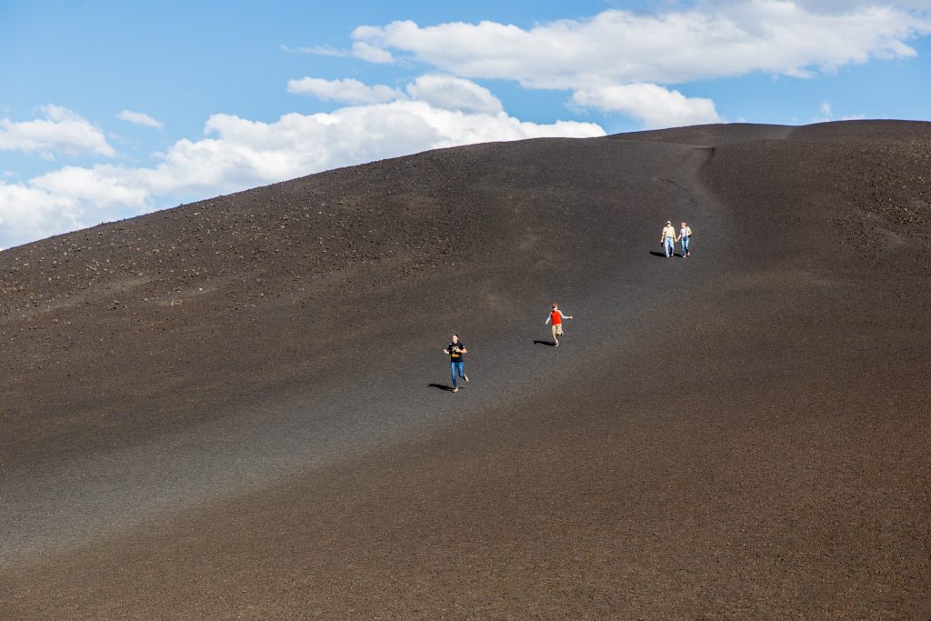 Vier wandelaars lopen omhoog over een steile helling van zwart vulkanisch gruis onder een blauwe lucht met wolken