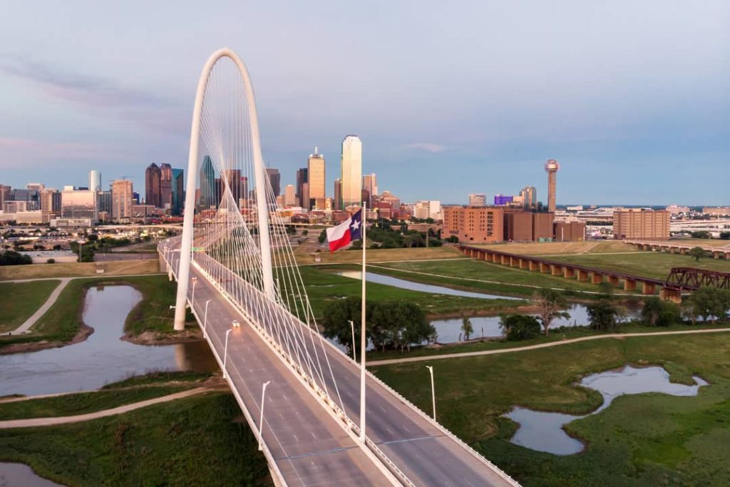 Witte boogbrug Margaret Hunt Hill Bridge over de Trinity River, met de skyline van Dallas en een Texaanse vlag