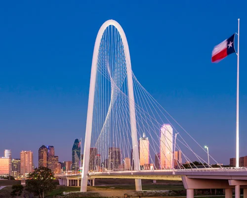 Witte boogbrug Margaret Hunt Hill Bridge bij schemering, met een wapperende Texaanse vlag en de skyline van Dallas