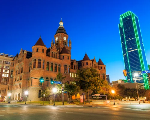 Verlicht rood bakstenen gerechtsgebouw met torentjes, met een groen omlijnde wolkenkrabber op de achtergrond bij nacht in Dallas