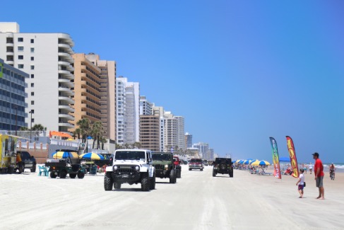Meerdere jeeps rijden over het brede witte zandstrand van Daytona Beach met hoge hotelgebouwen op de achtergrond