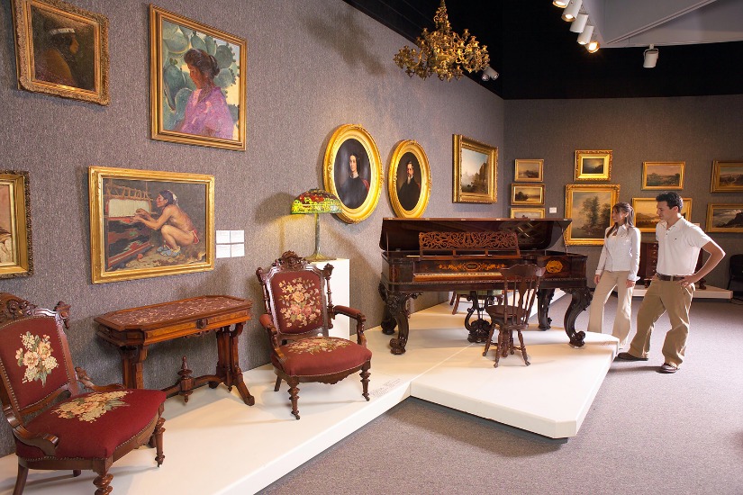 Bezoekers bekijken een zaal met gouden lijstschilderijen, antieke stoelen en een sierlijke piano in een museum in Daytona Beach