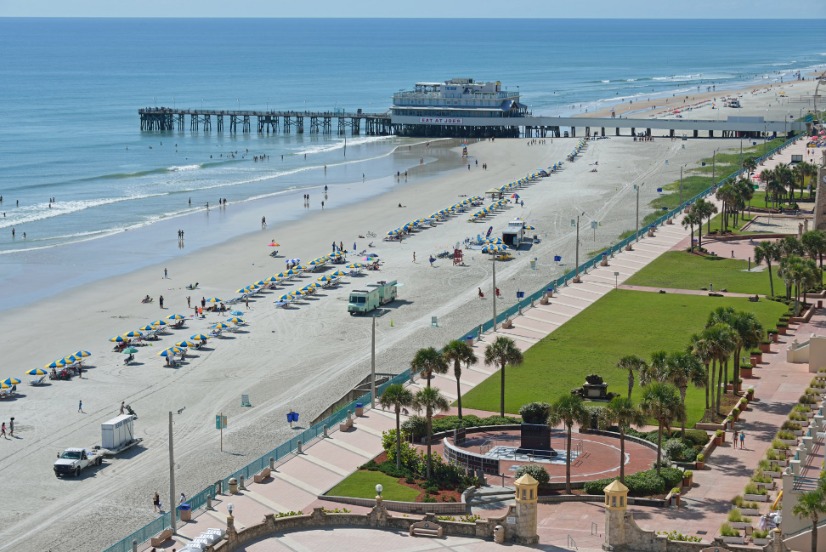 Luchtfoto van Daytona Beach in Florida met een lange pier, strandstoelen op het zand en een groene boulevard met palmbomen