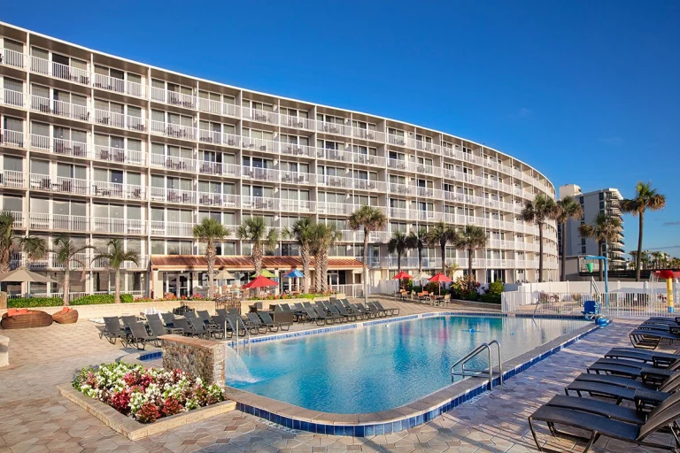 Groot zwembad omringd door ligstoelen en bloemperken voor een gebogen hotelgebouw in Daytona Beach