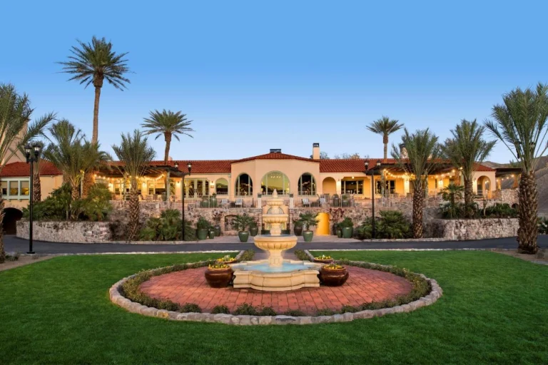 Stenen fontein op een groen gazon voor een resort met rode dakpannen en palmbomen tegen een woestijnberg in Death Valley