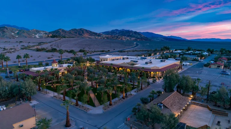 Verlicht resort met witte gebouwen en veel palmbomen omringd door woestijnbergen bij invallende avond in Death Valley