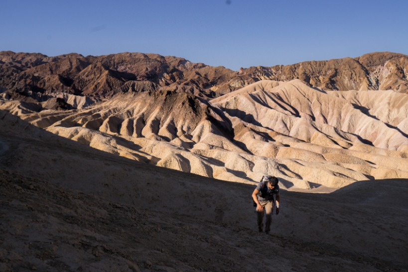 Wandelaar met rugzak beklimt een donkere heuvel met uitzicht op gegroefde, geel en bruin gestreepte badlandsformaties in Death Valley