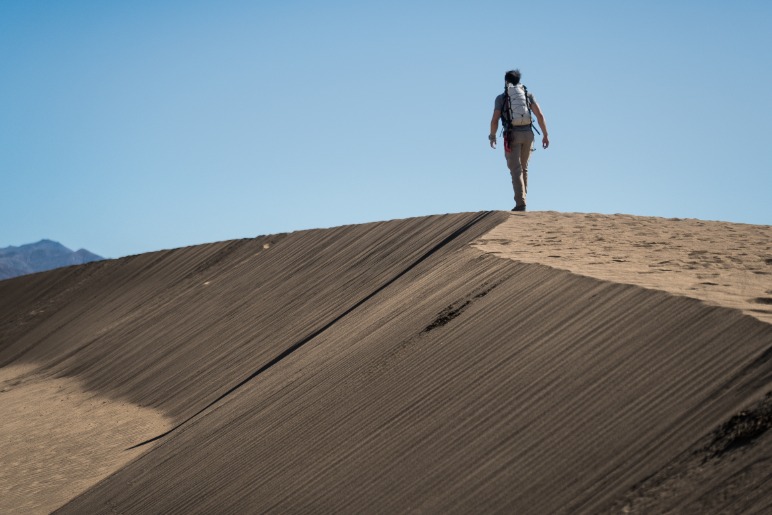Silhouet van een wandelaar met rugzak die over de scherpe kam van een zandduin loopt tegen een strakblauwe lucht