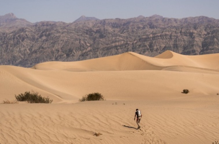 Eenzame wandelaar loopt over gegolfde zandduinen met kale bergen op de achtergrond in Death Valley National Park