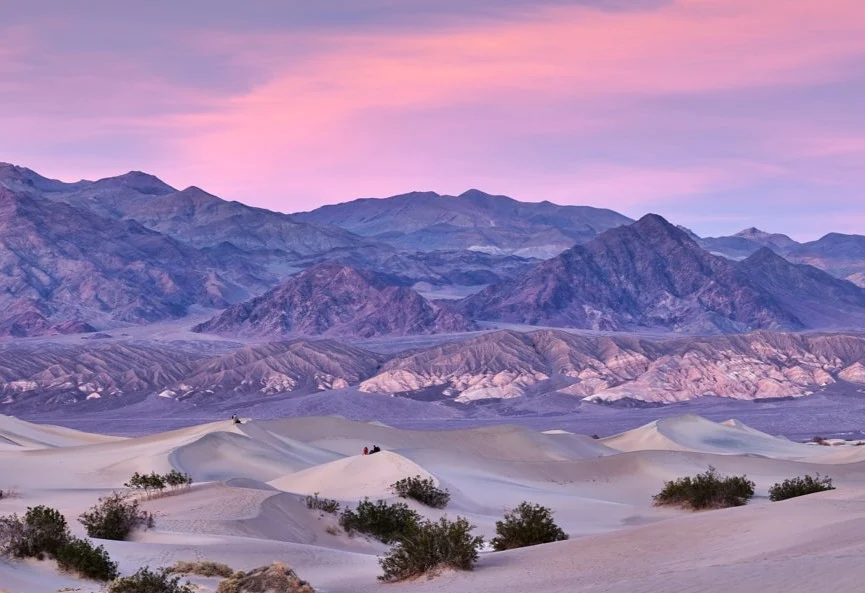 Golvende zandduinen kleuren zacht roze en paars onder een bewolkte avondlucht met bergen op de achtergrond in Death Valley